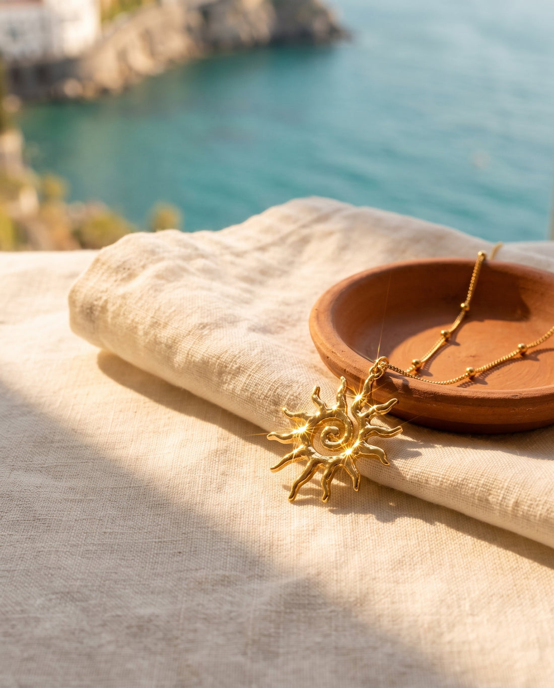 Gold sun pendant styled on cream linen with a terracotta dish and a sea view
