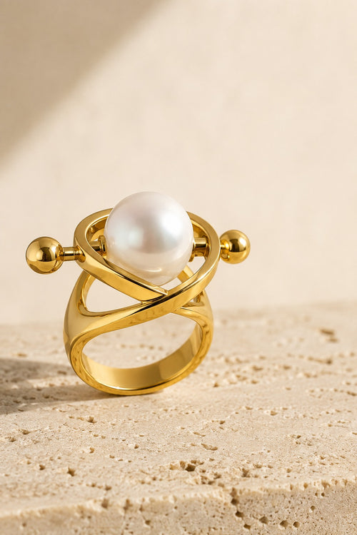 Atrani Orbit Ring standing on warm travertine stone, white pearl held inside crossing gold arcs