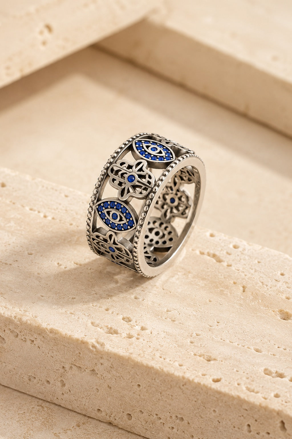 Cetara Evil Eye Ring on warm travertine stone, carved eye and hamsa band set with blue crystals